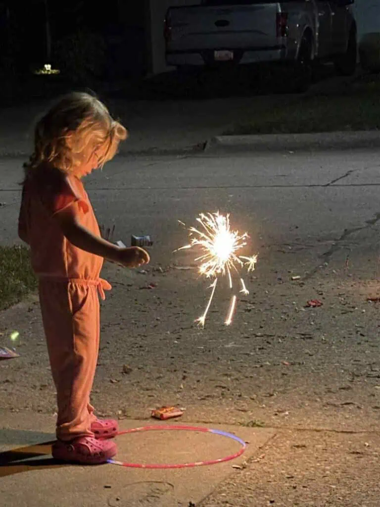 A young child in pink pajamas and Crocs holds a lit sparkler on a suburban street at night, celebrating Connor’s Big News amidst the quiet chaos of the neighborhood.
