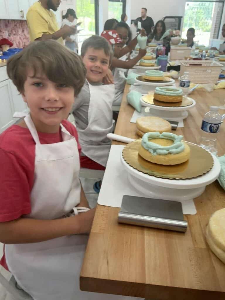Children wearing aprons sit at a table with small cakes and frosting tools, diving into a cake decorating activity that brings a dash of chaos and delight to the bright room, while adults watch in the background.