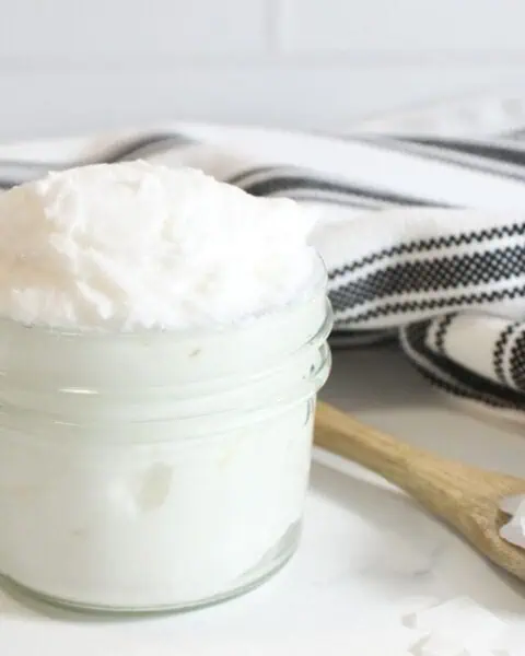 A small glass jar filled with whipped tallow sits next to a wooden spoon holding white flakes, with a black and white striped cloth in the background.