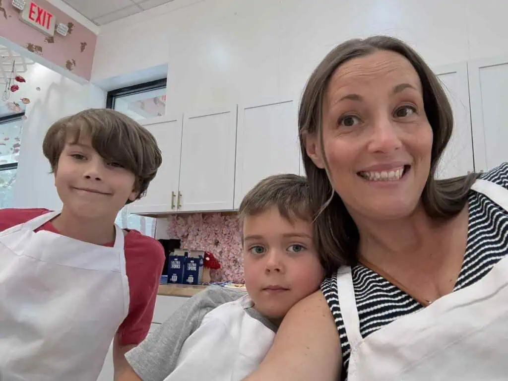 A woman and two young boys, all wearing aprons, pose for a selfie in a bright room with white cabinets and floral decor—amidst the cheerful chaos of baking a cake together.