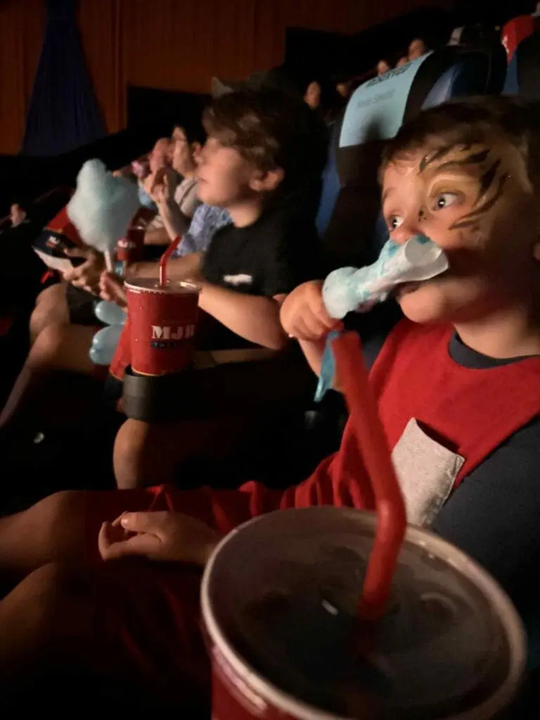 Children sit in a movie theater, one with face paint eating cotton candy and holding a drink with a red straw. Amid the chaos of excited whispers and laughter, other audience members are visible in the background.