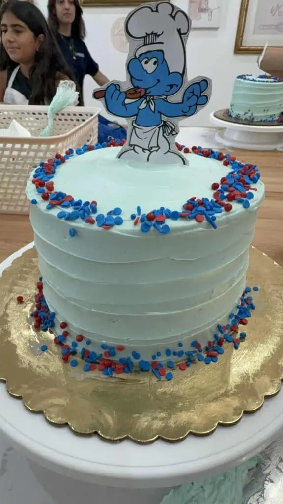 A light blue frosted cake with red and blue sprinkles is topped with a paper cutout of a cartoon chef Smurf, setting the stage for Connor’s Big News amid the cheerful chaos of several people in the background.