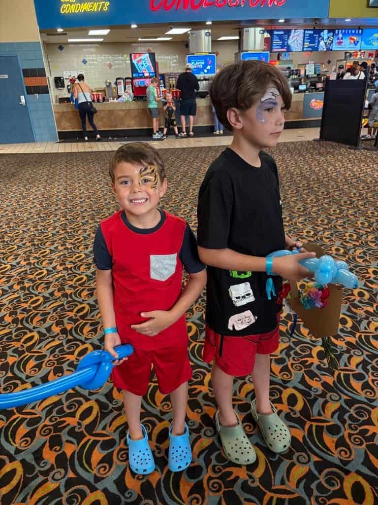 Two boys with face paint stand in a movie theater lobby. One smiles, holding a blue balloon like it’s Connor’s big news, while the other juggles balloons and toys amid the chaos of the busy concession stand and people in the background.