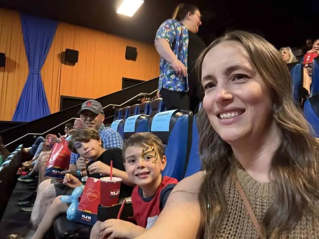 A woman and two children with snacks sit in a movie theater, with another adult behind them. Amid the happy chaos, one child sports face paint. Other people, eager for Connor's big news, are seated nearby.