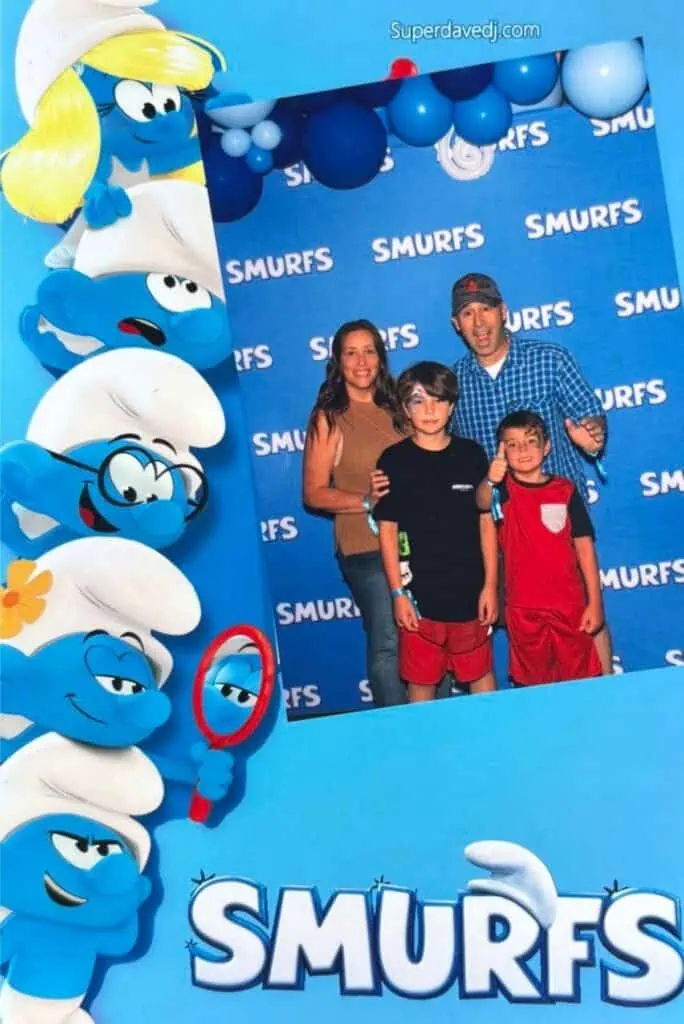 A family of four poses in front of a blue Smurfs-themed backdrop, surrounded by cartoon Smurf characters and the word "SMURFS" on the frame, ready to celebrate Connor’s Big News with cake and a touch of cheerful chaos.
