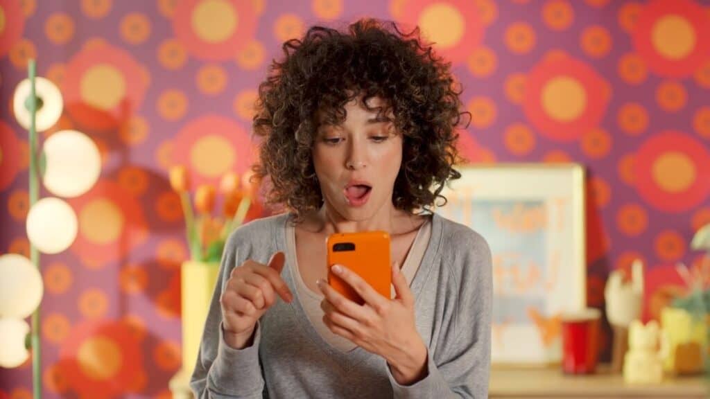 A woman with curly hair looks surprised at her orange smartphone, standing in front of a colorful, retro-patterned background—maybe she just read a Temu review.