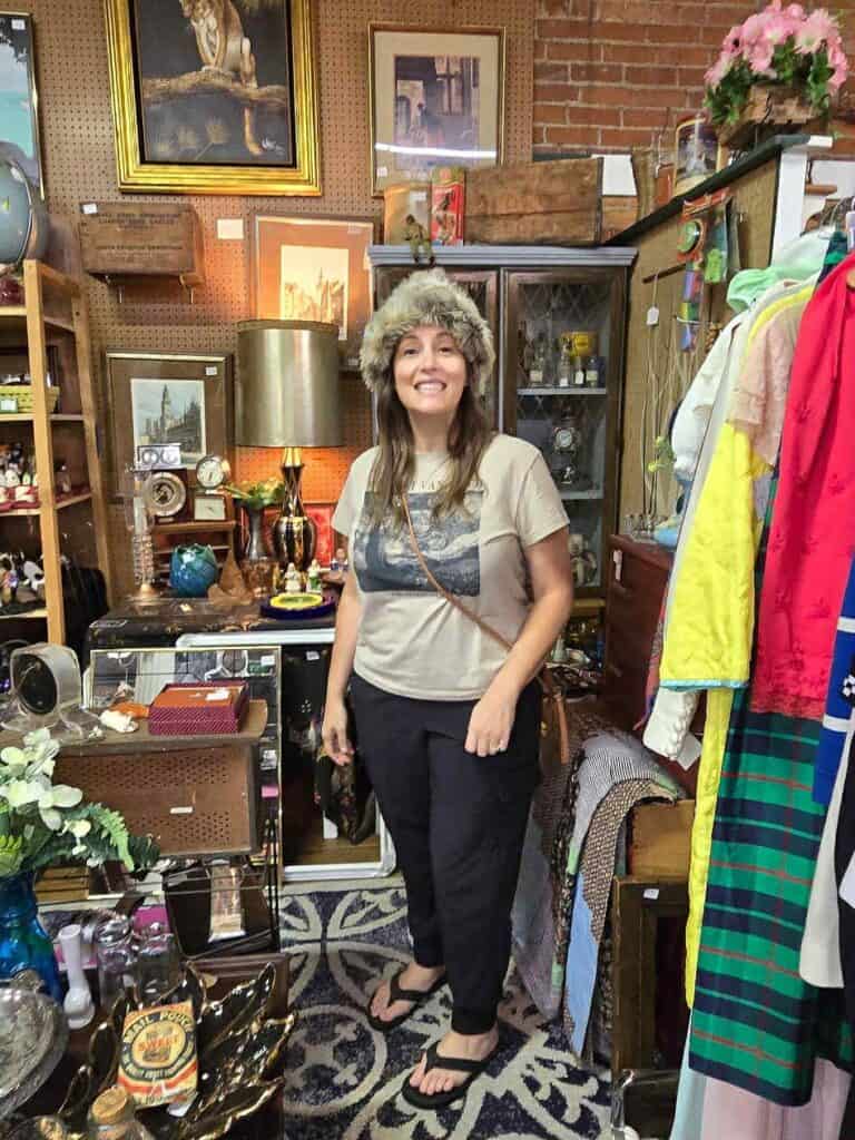 A woman wearing a faux fur hat, T-shirt, and sandals stands smiling in a vintage store surrounded by eclectic items and clothing, as if celebrating Connor’s Big News amid the delightful chaos of unique treasures.
