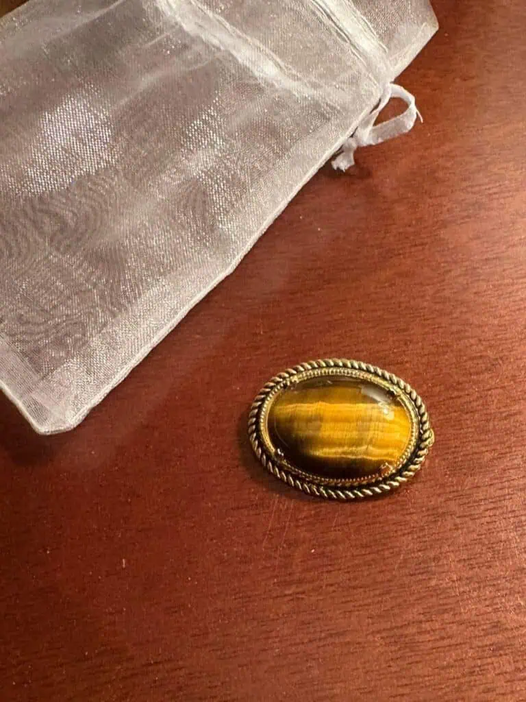 An oval tiger’s eye gemstone set in a decorative gold-colored metal frame sits on a wooden surface next to a sheer white drawstring pouch, capturing an elegant calm amid the chaos of Connor’s big news.