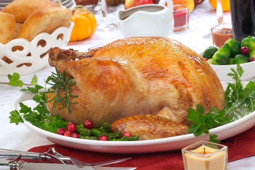 A whole roasted turkey garnished with herbs and cranberries on a white platter, set on a festive dining table with side dishes and drinks.