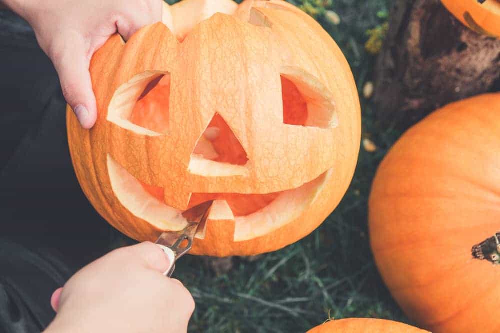 A person carves a smiling face into a pumpkin with a knife, creating a jack-o'-lantern. Other pumpkins are visible nearby on the grass.