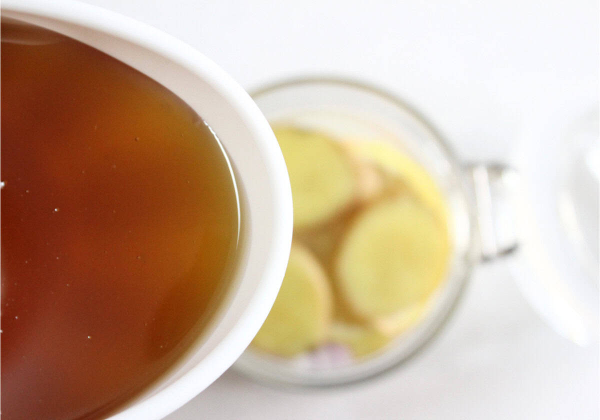 A close-up of brown homemade cough syrup in a white bowl next to a jar containing slices of ginger on a light surface.