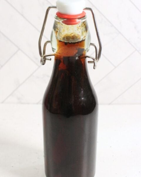 A clear glass bottle with a swing top lid is filled with dark brown diy vanilla extract and placed on a white surface against a white tiled background.