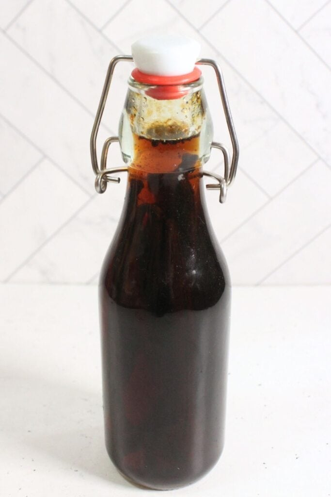 A clear glass bottle with a swing top lid is filled with dark brown diy vanilla extract and placed on a white surface against a white tiled background.