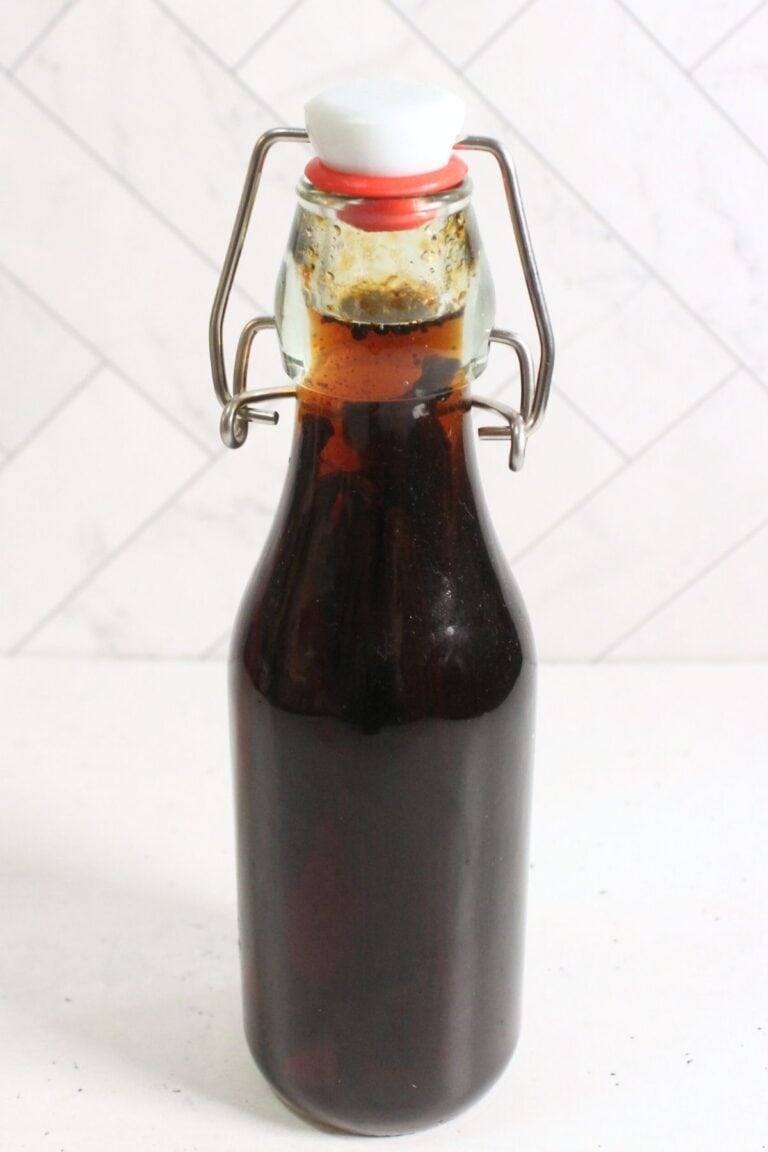 A clear glass bottle with a swing top lid is filled with dark brown diy vanilla extract and placed on a white surface against a white tiled background.