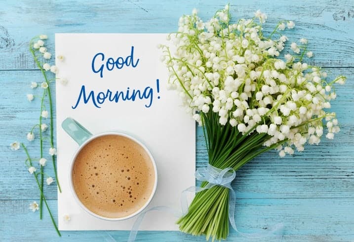 A cup of coffee, a bouquet of white flowers, and a card with "Good Morning!" written on it are arranged on a light blue wooden surface.