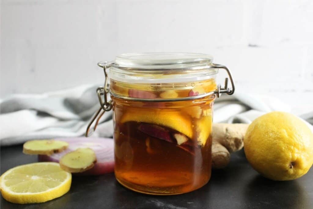 A glass jar filled with homemade cough syrup—honey, lemon slices, and ginger—sits on a dark surface with whole and sliced lemon and ginger beside it.
