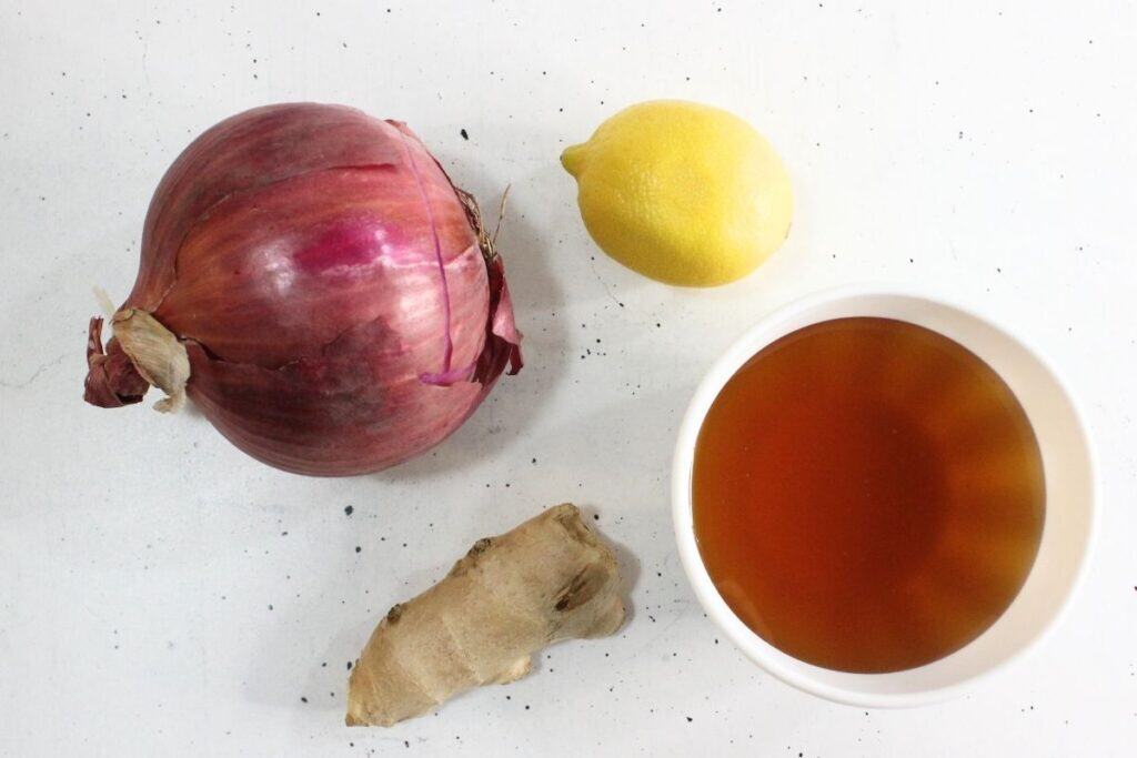 A red onion, a lemon, a piece of ginger root, and a bowl of brown liquid—classic ingredients for homemade cough syrup—are arranged on a white surface.