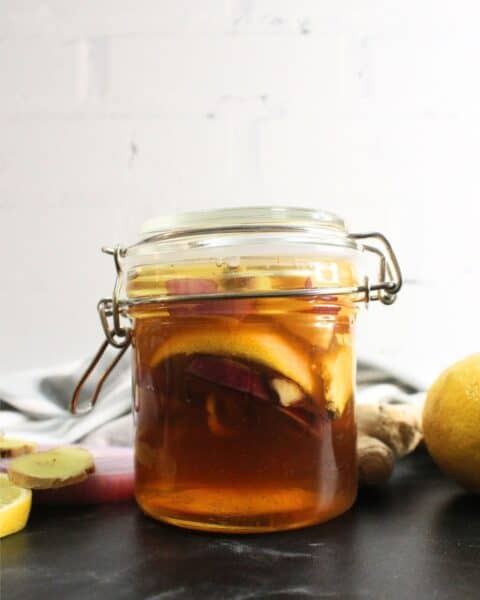 A glass jar with a hinged lid containing homemade cough syrup made from honey, lemon slices, and ginger, placed on a dark surface with fresh lemon and ginger nearby.