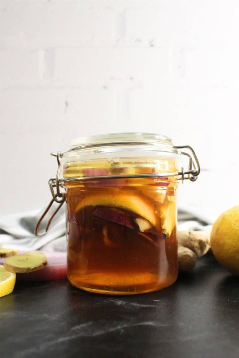 A glass jar with a hinged lid containing homemade cough syrup made from honey, lemon slices, and ginger, placed on a dark surface with fresh lemon and ginger nearby.