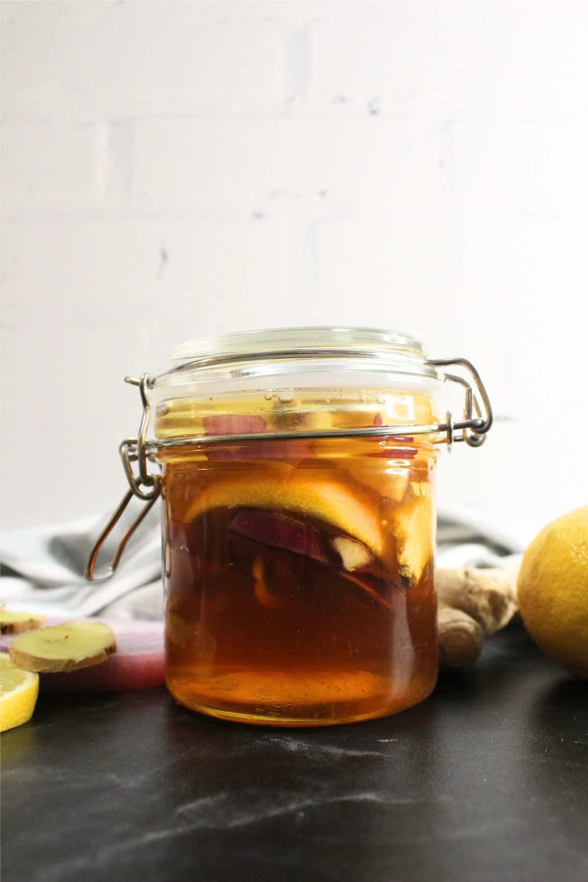 A glass jar with a hinged lid containing homemade cough syrup made from honey, lemon slices, and ginger, placed on a dark surface with fresh lemon and ginger nearby.