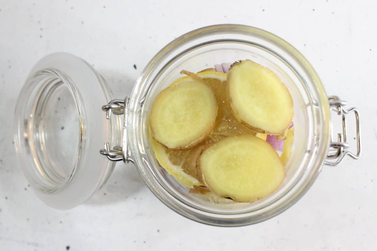 A glass jar with the lid open containing lemon slices, ginger slices, and what appears to be homemade cough syrup on a white surface.