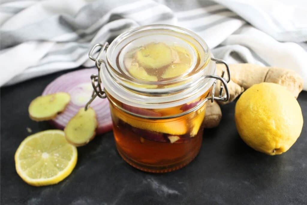 A glass jar with a metal clasp filled with homemade cough syrup—a honey mixture containing lemon, ginger, and red onion slices—surrounded by fresh lemon, ginger, and onion on a dark surface.