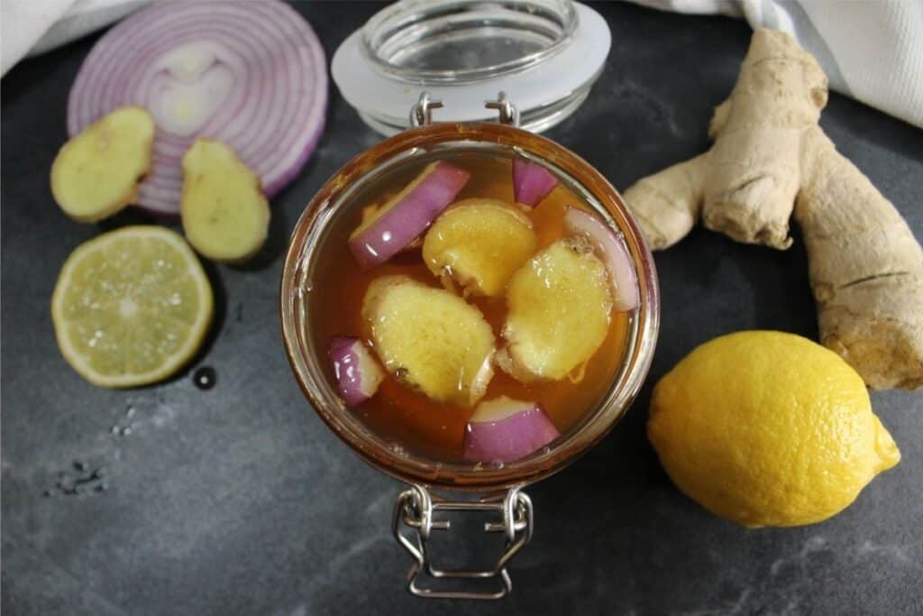 A jar of homemade cough syrup with sliced ginger and red onion in liquid, surrounded by a lemon, lime half, ginger root, and a slice of red onion on a dark surface.