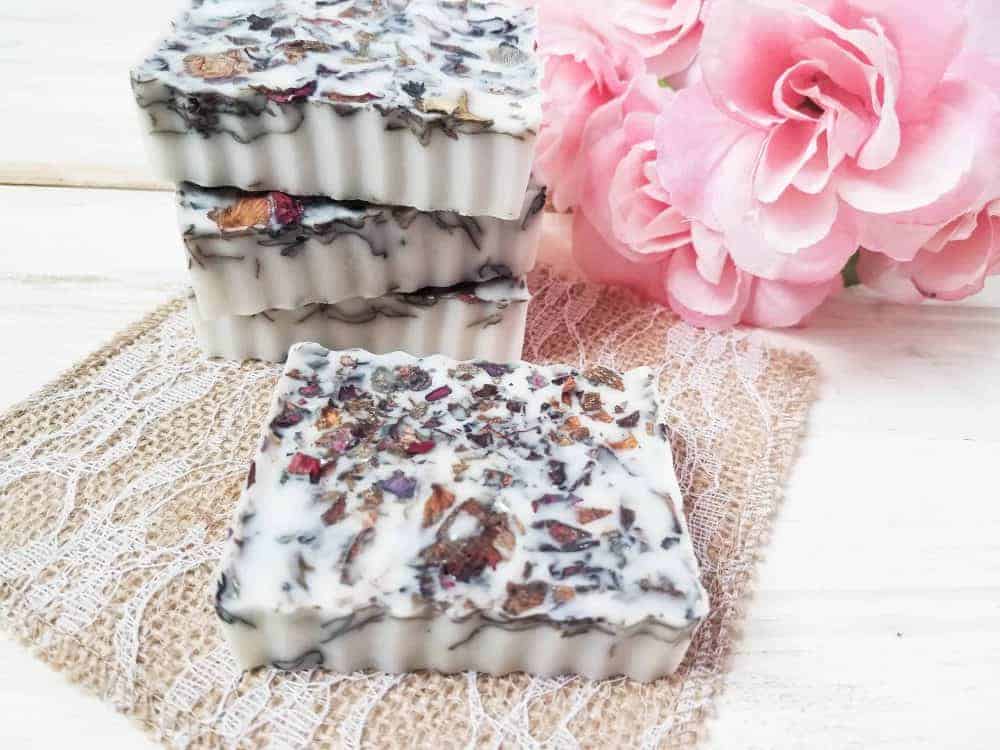 Four handmade floral soaps crafted with DIY natural products and visible dried flower petals are stacked next to pink artificial roses on lace and burlap fabric.