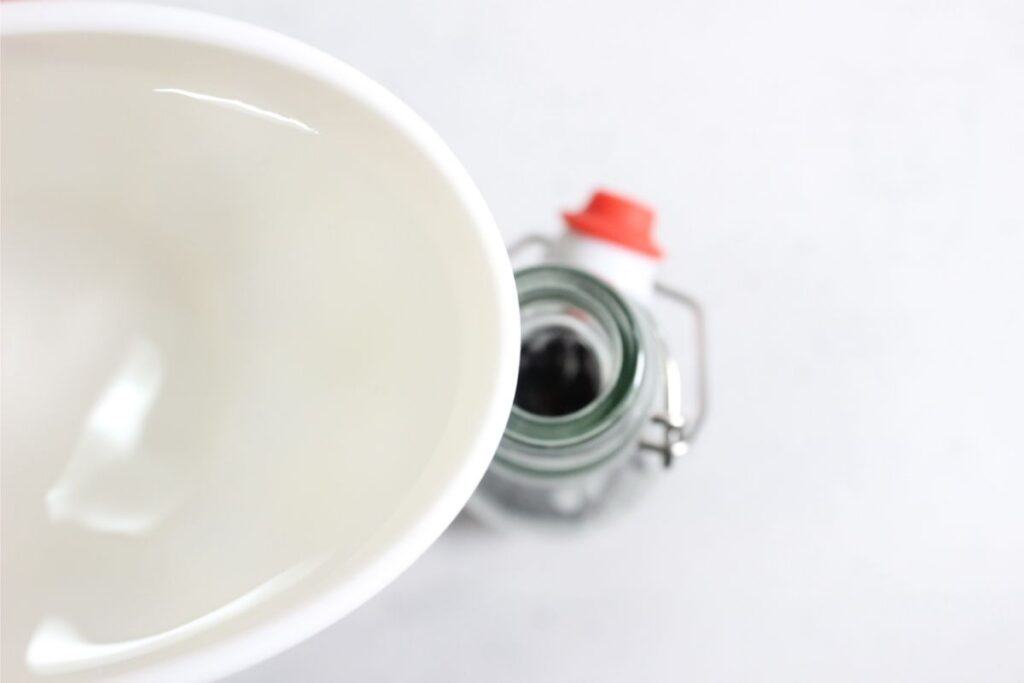A white bowl with liquid, perfect for making DIY vanilla extract, is being positioned above a glass bottle with a red and white swing top cap.