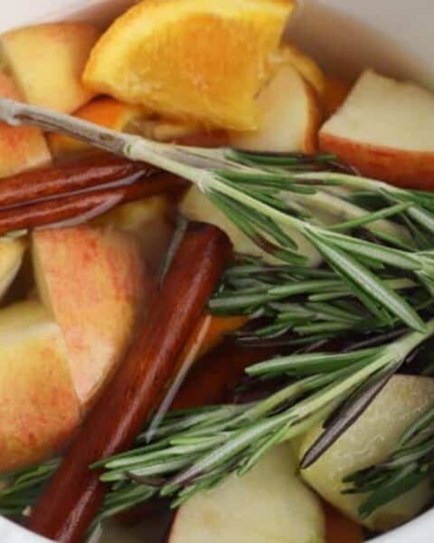 Sliced apples, orange wedges, cinnamon sticks, and rosemary sprigs in a white pot filled with water make a cozy Autumn simmer pot—perfect for those who love autumn recipes and fragrant simmer pot recipes.