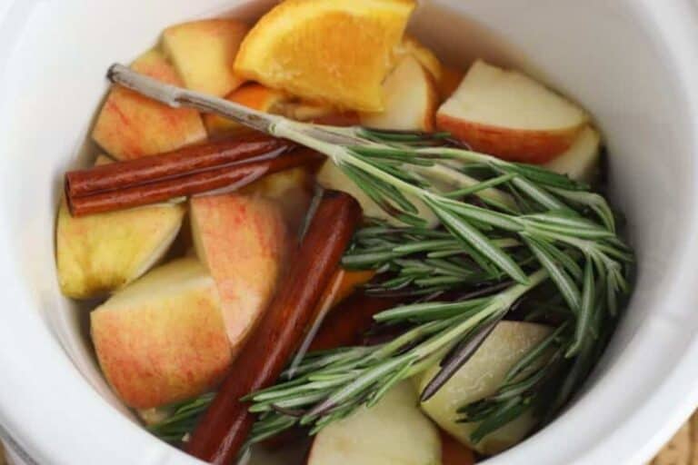 Sliced apples, orange wedges, cinnamon sticks, and rosemary sprigs in a white pot filled with water make a cozy Autumn simmer pot—perfect for those who love autumn recipes and fragrant simmer pot recipes.