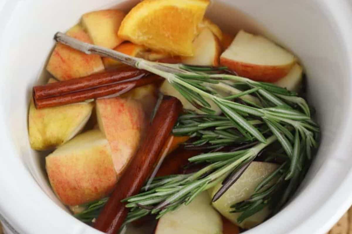 Sliced apples, orange wedges, cinnamon sticks, and rosemary sprigs in a white pot filled with water make a cozy Autumn simmer pot—perfect for those who love autumn recipes and fragrant simmer pot recipes.
