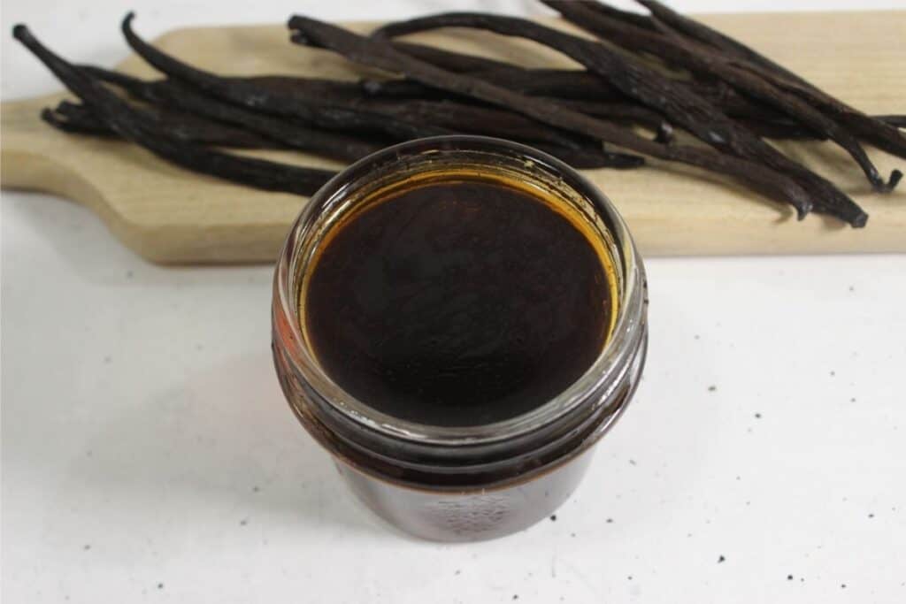 A glass jar filled with dark liquid sits on a white surface, with dried vanilla beans on a wooden board in the background—perfect for making your own DIY vanilla extract.
