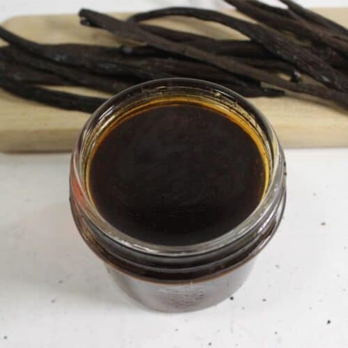 A glass jar filled with dark liquid sits on a white surface, with dried vanilla beans on a wooden board in the background—perfect for making your own DIY vanilla extract.