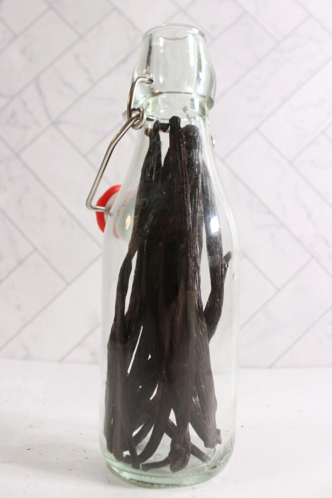 A clear glass bottle filled with whole dried vanilla beans, perfect for diy vanilla extract, sealed with a metal clasp lid and placed on a white surface with a tiled background.