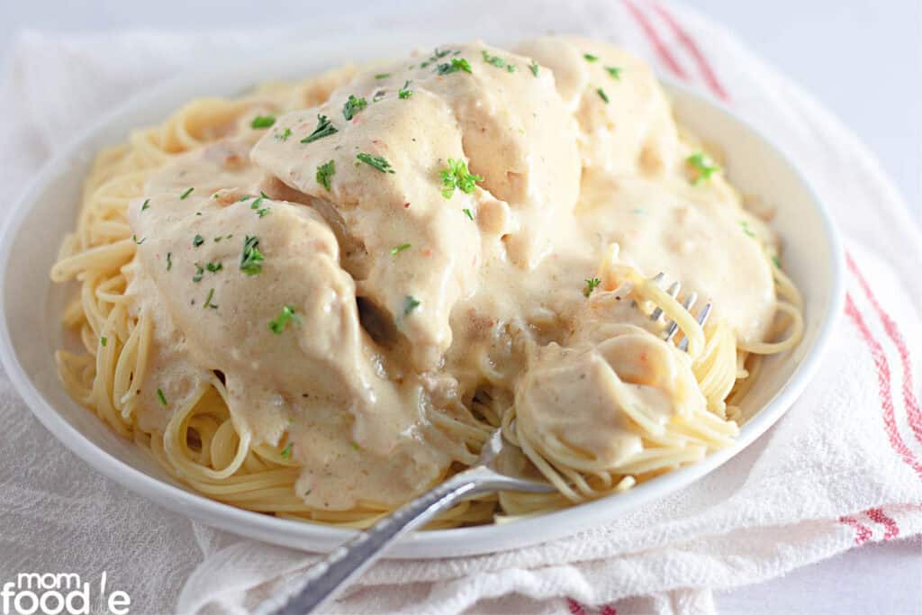 A white bowl filled with spaghetti topped with creamy slow cooker chicken pieces and garnished with chopped parsley, placed on a white cloth with red stripes.