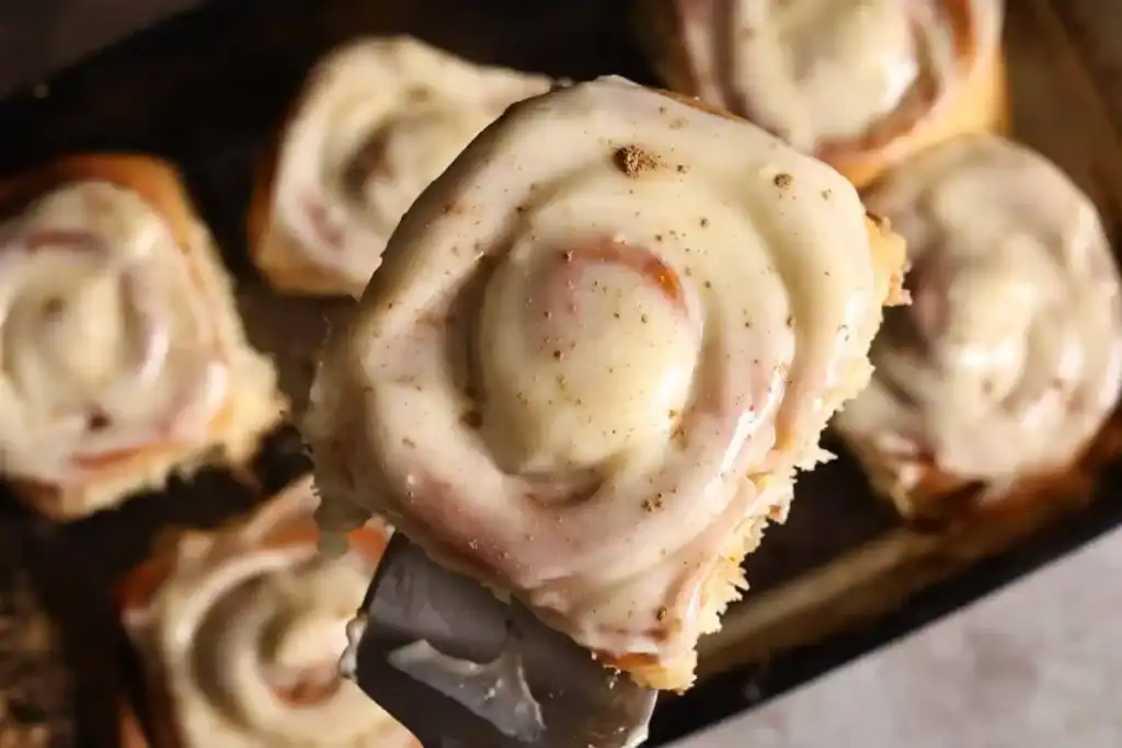 A close-up of a cinnamon roll topped with icing and apples being lifted from a tray of other cinnamon rolls—one of our favorite creative apple uses.