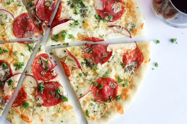 A sliced pizza topped with pepperoni, red onion, herbs, and melted cheese is arranged on a white surface—try adding thin apple slices for a twist inspired by creative uses for apples—with a glass of dark beverage partially visible in the corner.