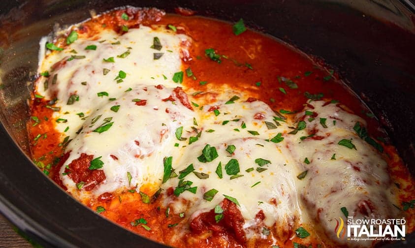Four chicken breasts topped with melted cheese and tomato sauce in a slow cooker, garnished with chopped parsley—perfect for easy dinners or tasty Crockpot dinners.