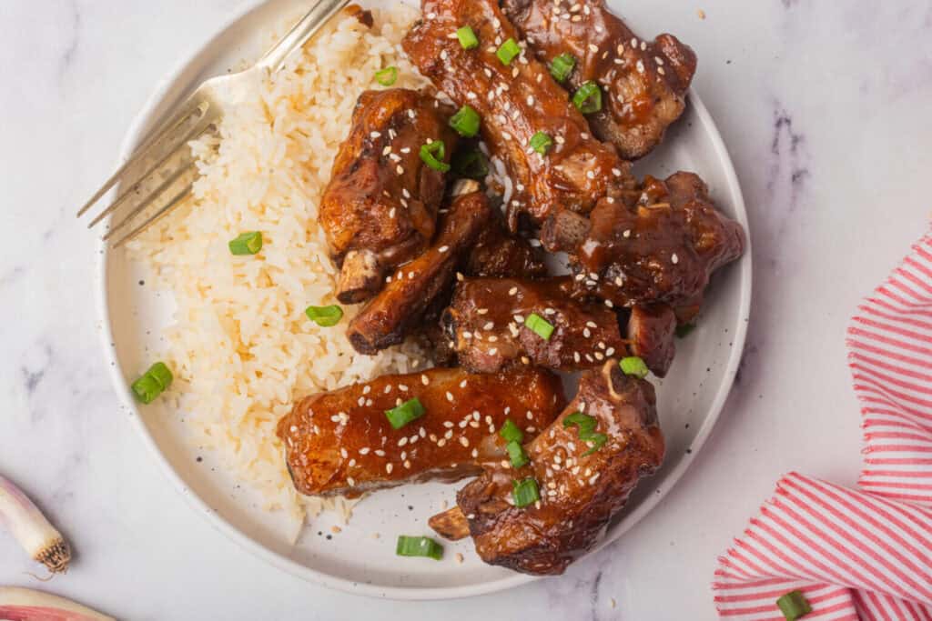 A plate of white rice and glazed pork ribs garnished with sesame seeds and chopped green onions, with a fork on the side—perfect for easy meals or as a delicious addition to your favorite slow cooker recipes.