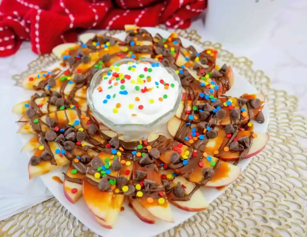 A plate of sliced apples, perfect for creative apple uses, topped with chocolate, caramel, sprinkles, and chocolate chips, arranged around a central bowl of whipped cream.