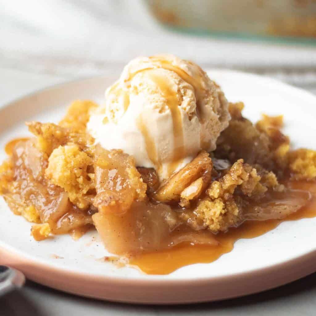 A serving of apple cobbler—one of the tastiest apple recipes—topped with vanilla ice cream and drizzled with caramel sauce on a white plate.