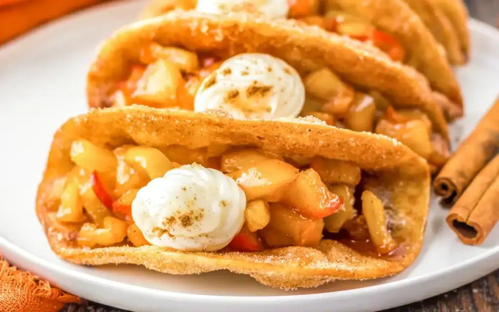 Close-up of dessert tacos filled with diced apples—an example of creative apple uses—topped with whipped cream, set on a white plate with cinnamon sticks nearby.