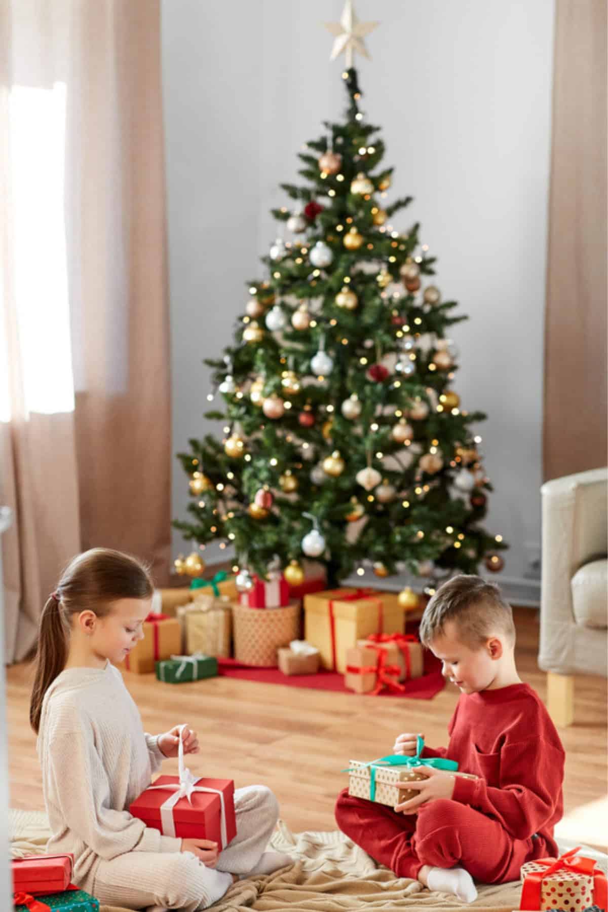 Two children in pajamas sit on the floor opening presents in front of a decorated Christmas tree with wrapped gifts underneath. Discover how to get a Free Christmas Tree with our helpful Free Christmas Tree guide.