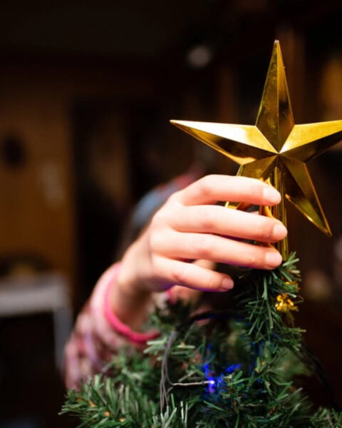 A person places a gold star ornament on top of a decorated artificial Christmas tree indoors, showcasing festive spirit and offering inspiration for those wondering how to get a Free Christmas Tree this holiday season.
