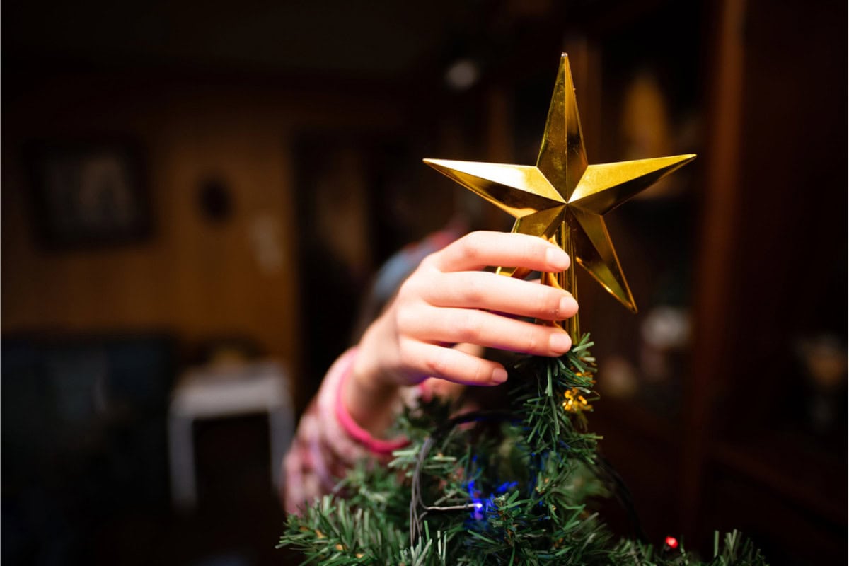 A person places a gold star ornament on top of a decorated artificial Christmas tree indoors, showcasing festive spirit and offering inspiration for those wondering how to get a Free Christmas Tree this holiday season.