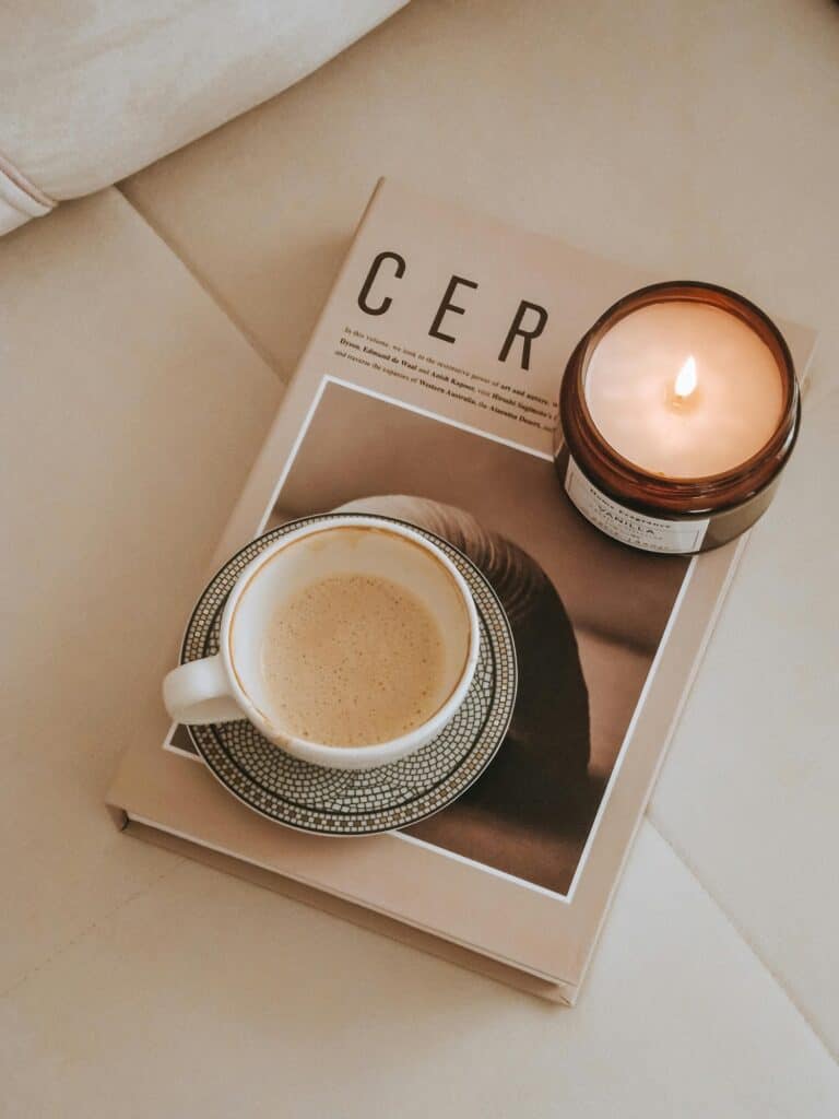 A cup of coffee on a saucer, a lit candle, and a magazine are arranged on a beige surface.