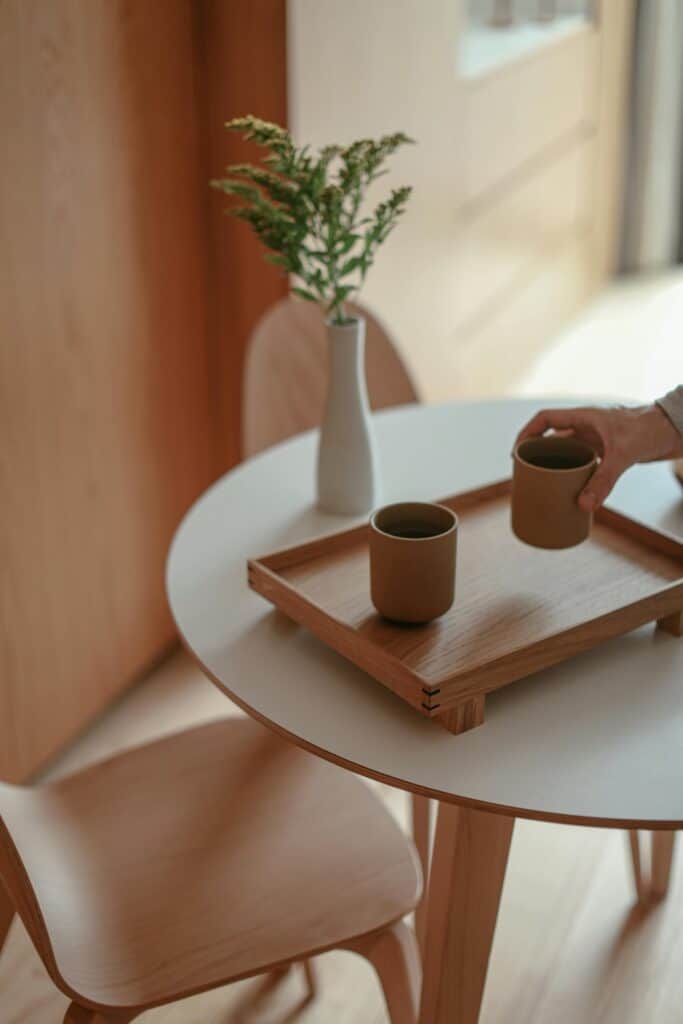 A round white table with a wooden tray holding two cups, a hand reaching for one cup, and a white vase with greenery in the background.