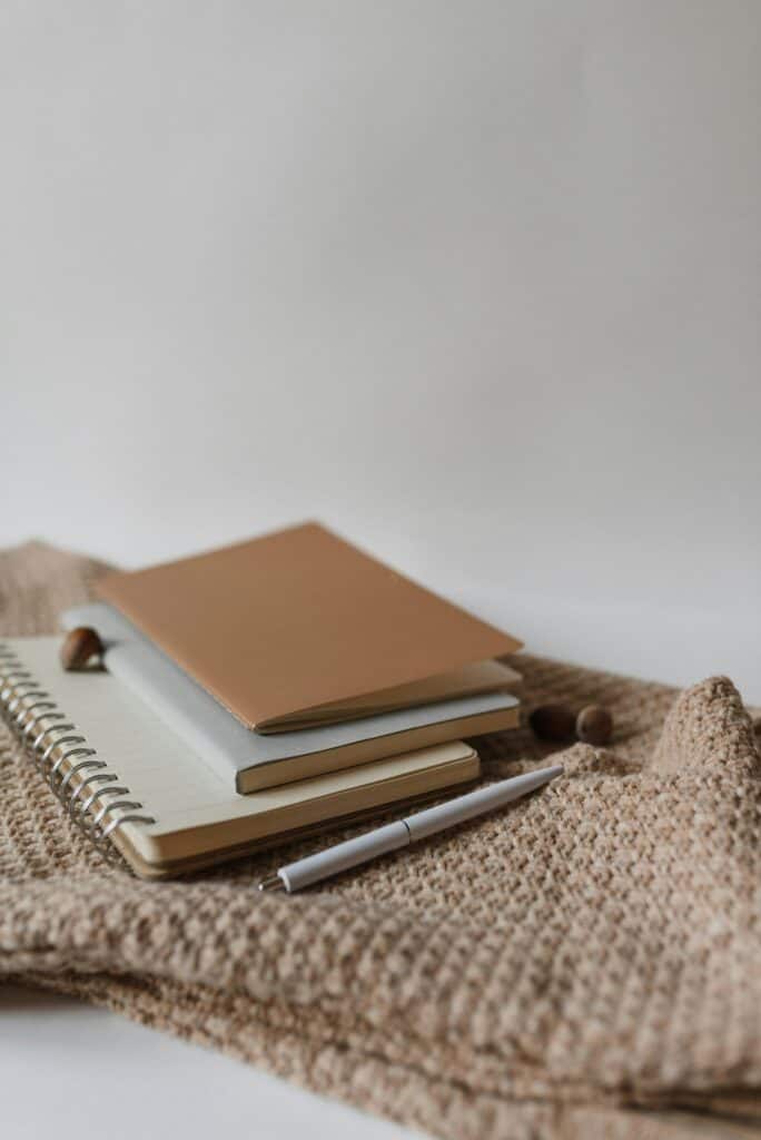 A brown notebook, a gray book, and a spiral-bound notepad are stacked on a textured beige fabric, with a silver pen and small round objects nearby.