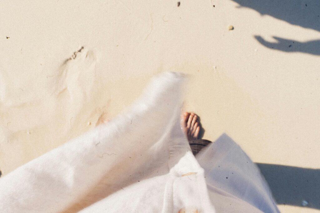 A person wearing a light-colored garment stands barefoot on sandy ground, with part of their foot and shadow visible.